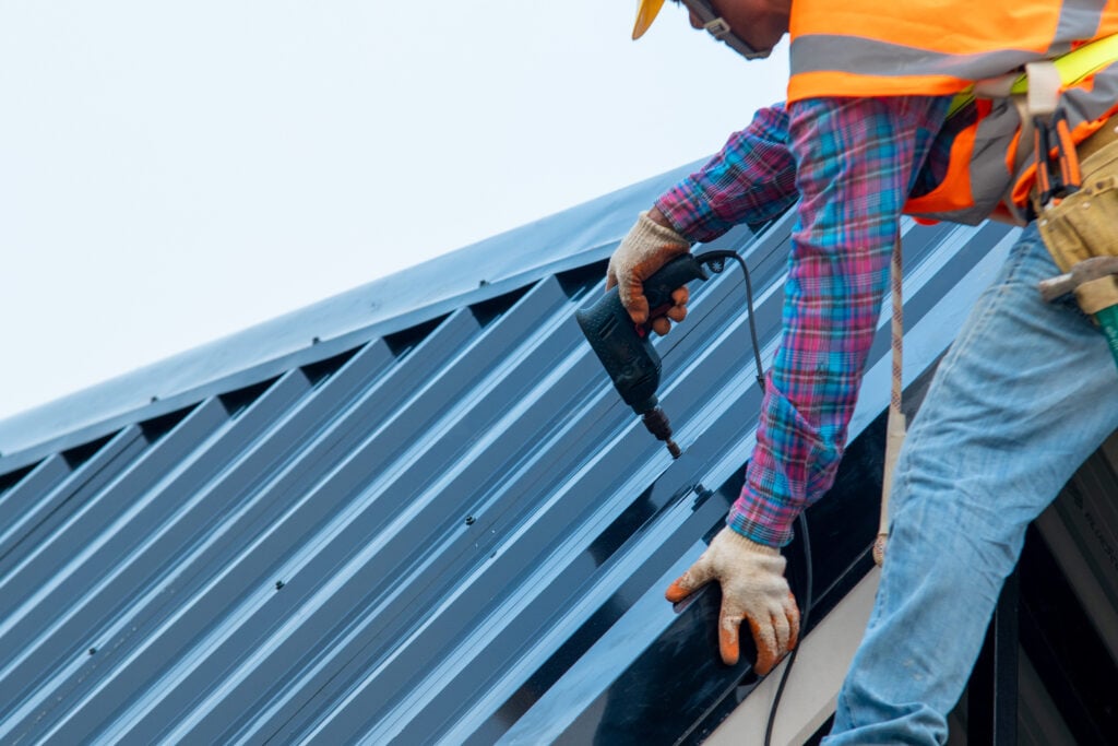 Construction worker wearing safety harness belt during working on roof structure of building on construction site,Roofer using air or pneumatic nail gun and installing metal roof tile on top new roof.