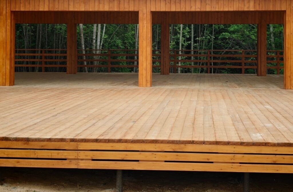 Open wooden pavilion structure with a sloped metal roof, surrounded by trees, featuring a spacious wooden deck