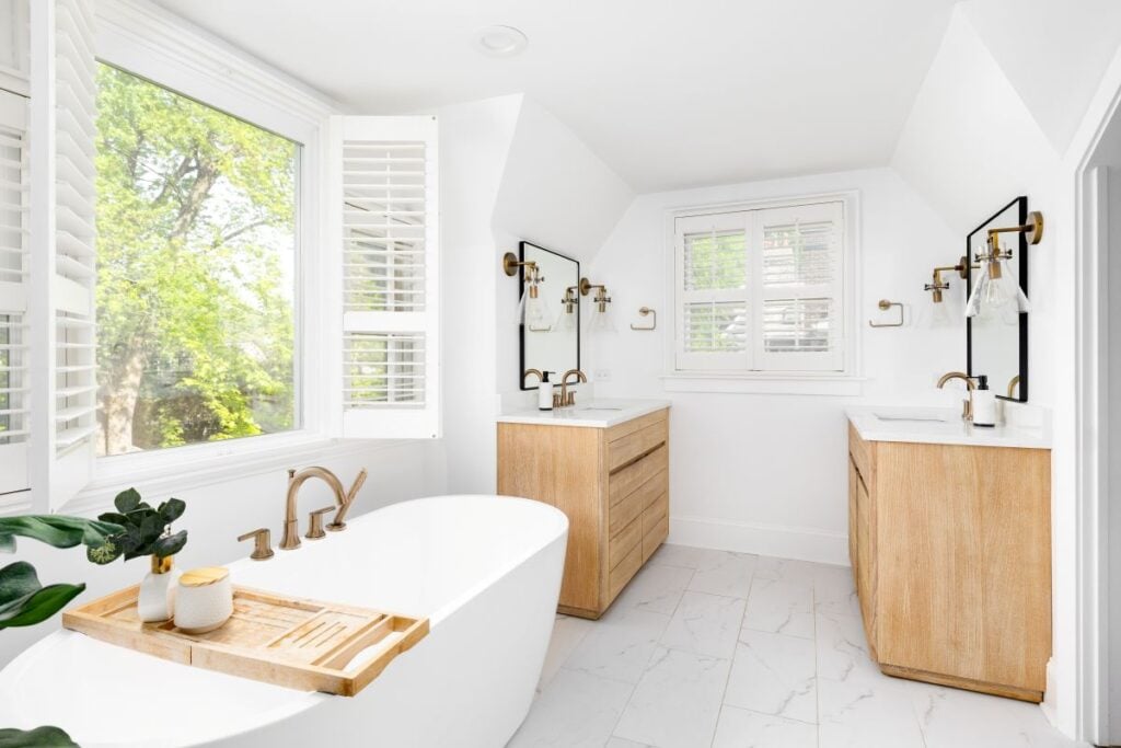 A beautiful bathroom with a freestanding soaking tub with a gold faucet, natural wood vanity cabinets, marble tile flooring, and bronze fixtures.