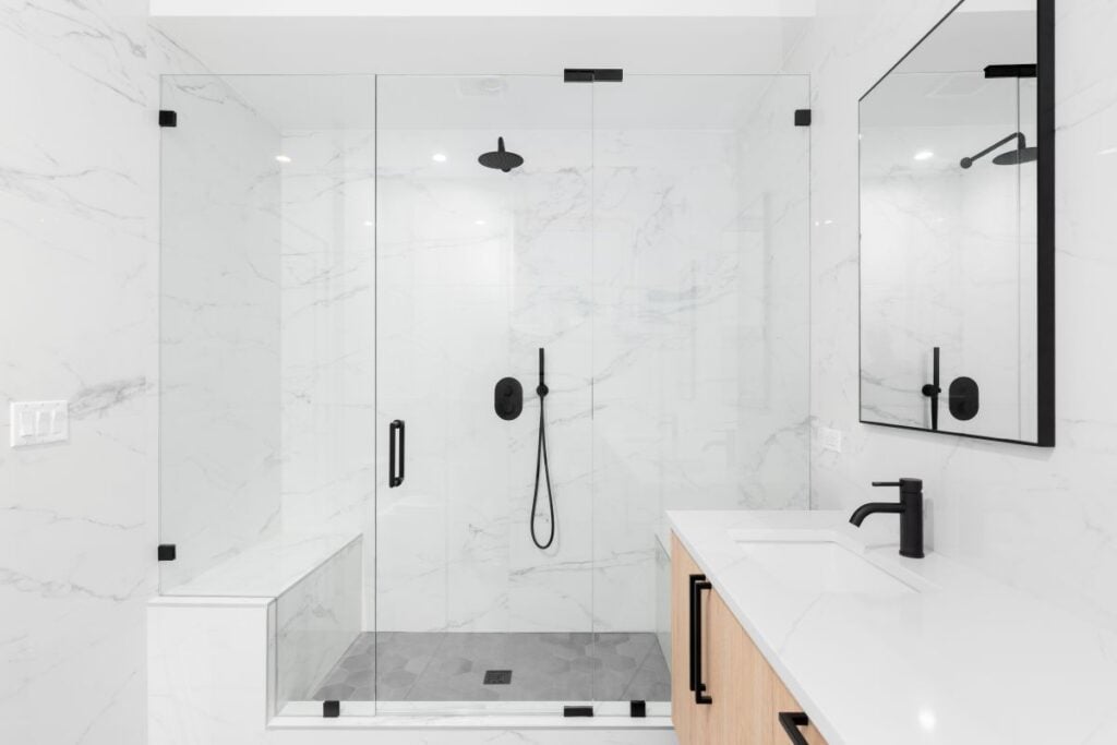 A beautiful luxury, modern bathroom with a light wood cabinet, walk-in shower with marble tiled walls, and black faucets and hardware.
