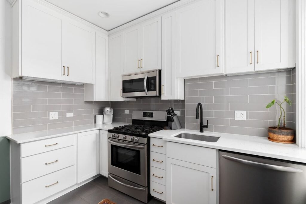 A small, white kitchen with a grey subway tile backsplash, stainless steel appliances, and grey tiled floor. No brands or labels.
