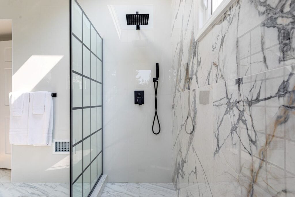 Luxury modern walk-in shower with marble walls, black fixtures and glass partition in bright bathroom
